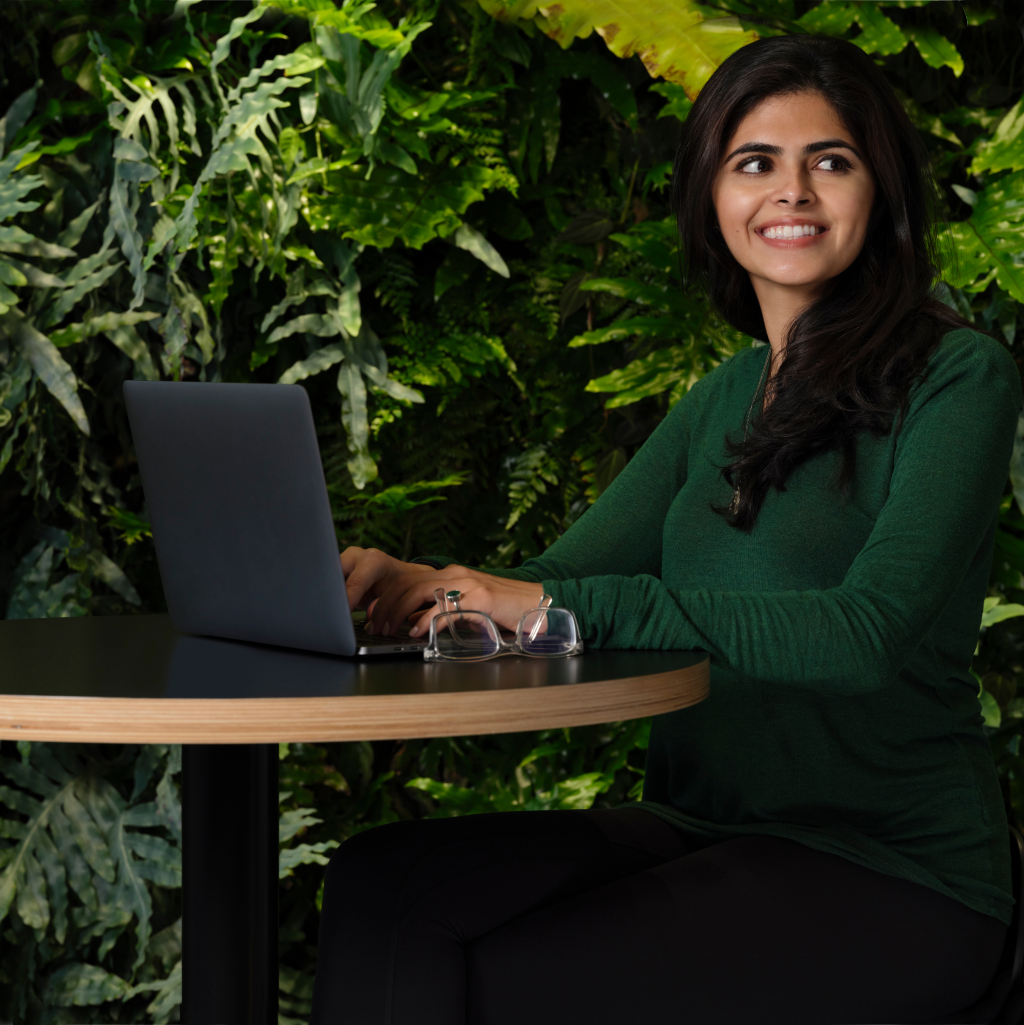 A woman in a green shirt sits at a round wooden‑top table with a laptop and glasses, smiling in front of a lush green plant wall.